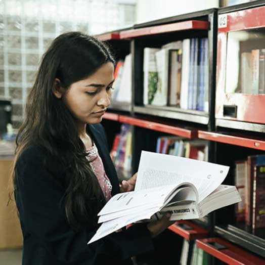 Student reading a book while standing near library bookshelves at KIM Harihar library