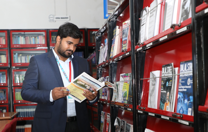 Student reading a book in the library with bookshelves in the background at Kirloskar Institute of Management