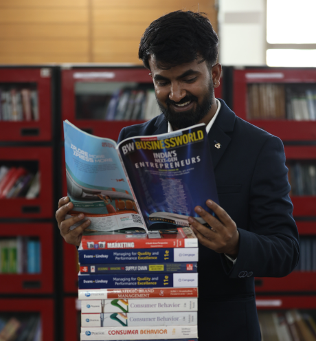 Student in library at KIM reading a business magazine while holding a stack of management books