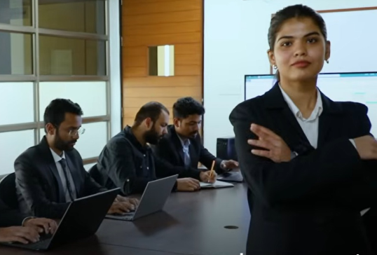Student in formal attire standing confidently with team working on laptops in background, representing Analytics and AI focus